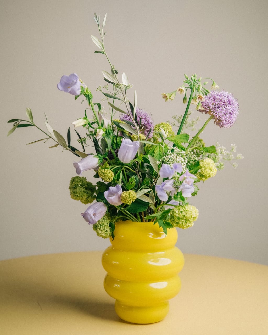 Gelbe Wellenvase mit Glockenblumen, Zierlauch, Schneeball und Olivenzweigen auf einem gelben Tisch