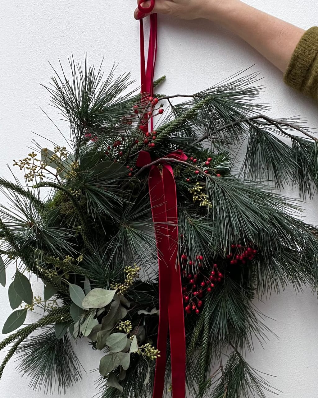 Ella hält einen Kranz aus Kiefer, Eukalyptus und roten Beeren am roten Samtband vor eine weiße Wand