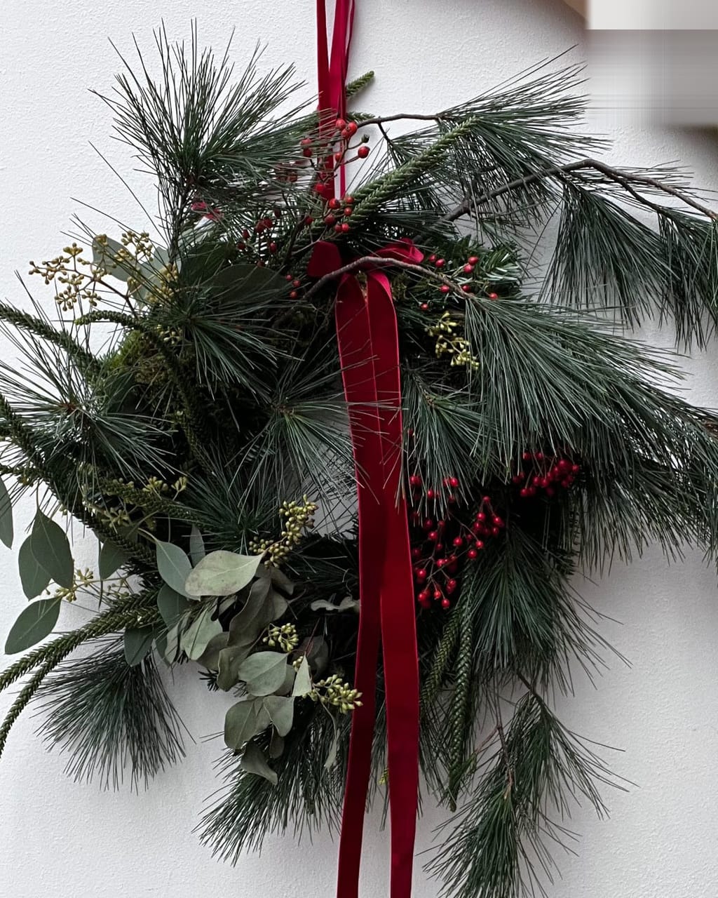 Kranz aus Kiefer, Eukalyptus und roten Beeren mit rotem Samtband, hängend vor einer weißen Wand
