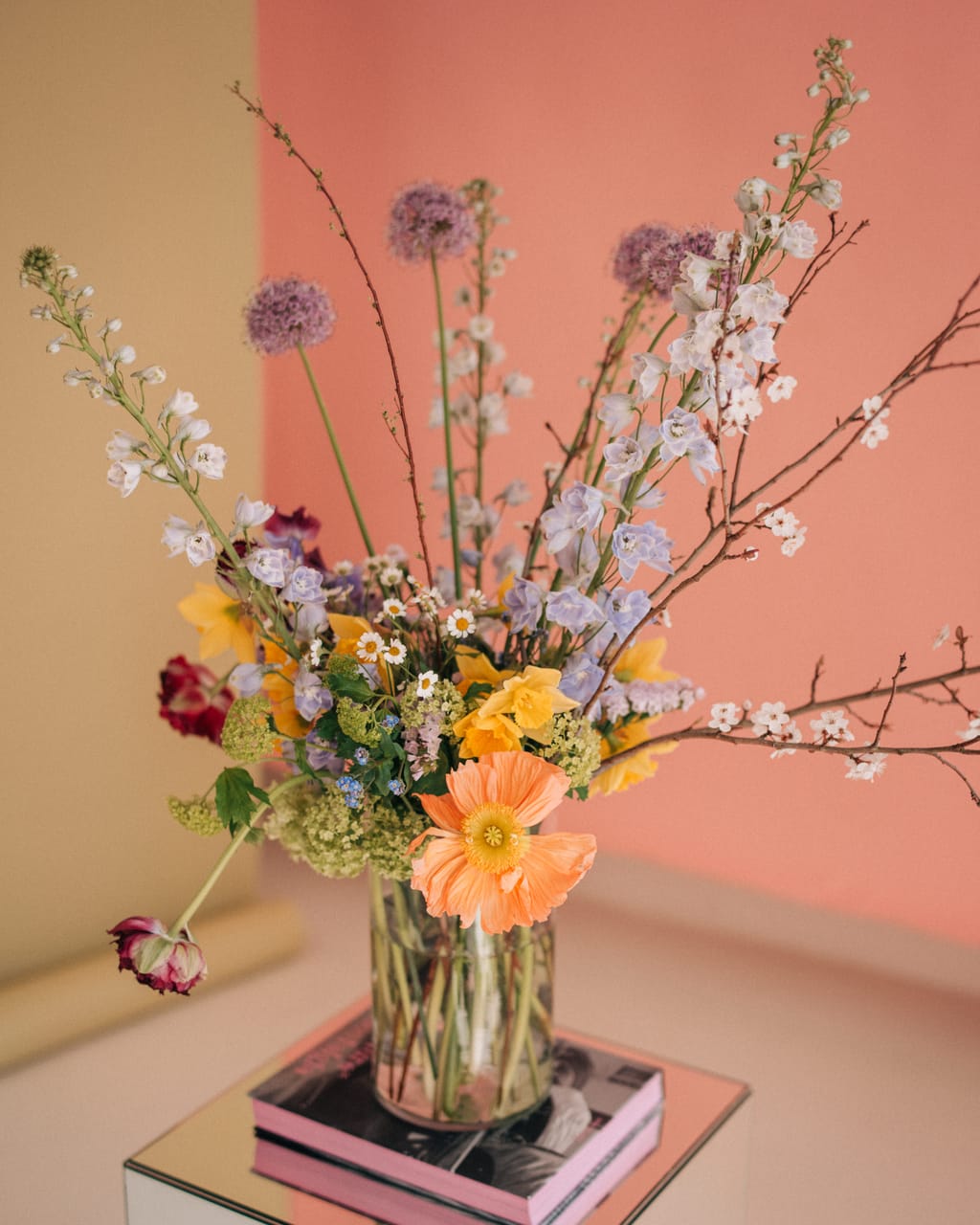 Frühlingsstrauß mit Zierlauch, Rittersporn, Narzissen, Islandmohn und Kirschblütenzweigen in einer Glasvase auf einem Spiegelsockel vor einer rosa Wand