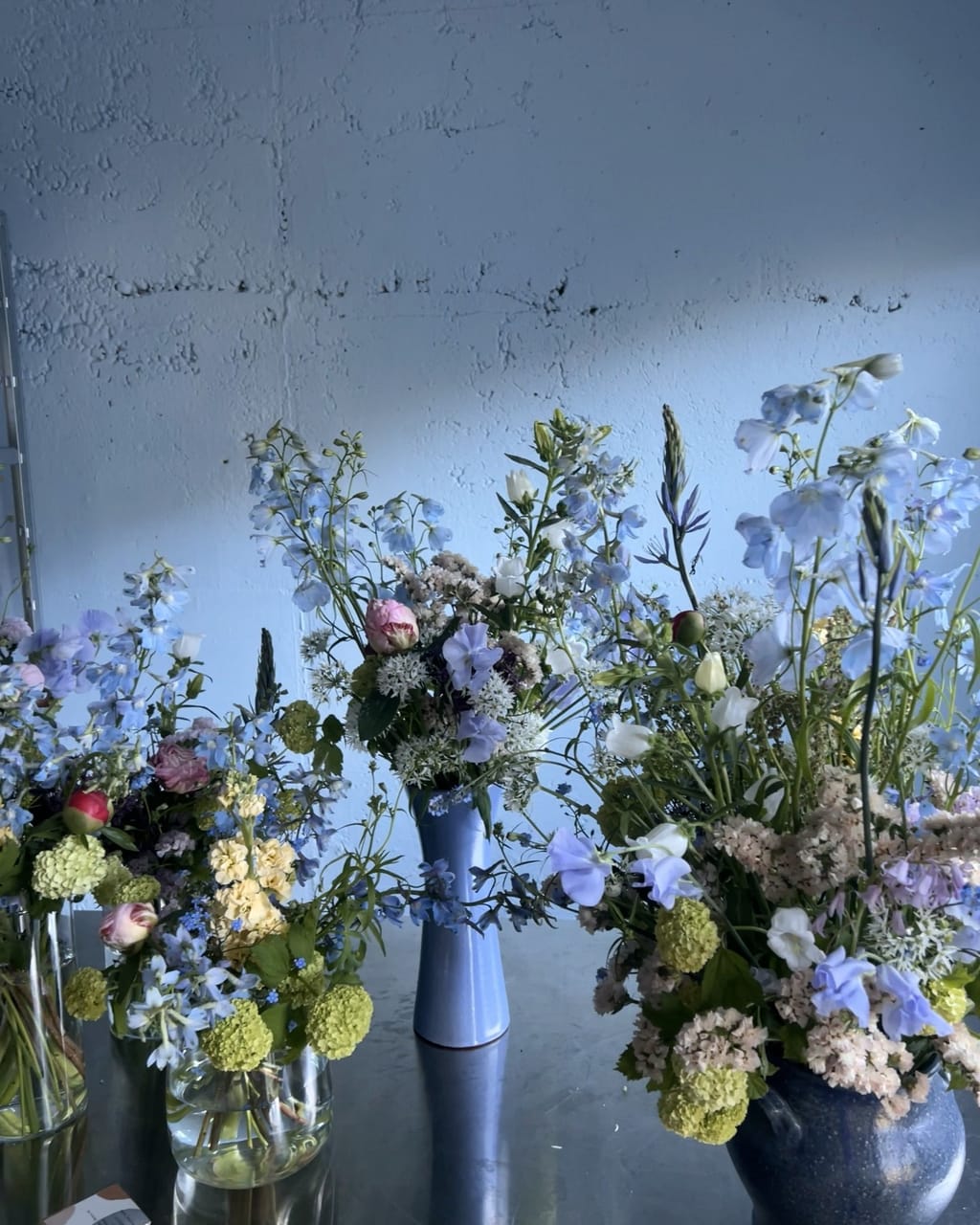 Blaue Keramikvasen mit Rittersporn, Wicken und Schneeball vor einer grauen Wand